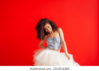 Charming mixed race girl smiling and posing in denim top and tulle tutu skirt. Attractive laughing african woman with glitter on her face having fun on camera in studio. Red background.