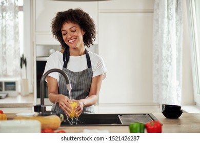 Charming mixed race woman in apron standing in kitchen ad washing yellow paprika in sink. On kitchen counter are different sorts of vegetables. - Powered by Shutterstock