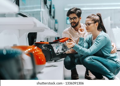 Charming Multicultural Couple Crouching And Looking At Vacuum Cleaner They Want To Buy. Tech Store Interior.