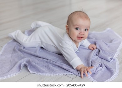 Charming Newborn Baby In A White Bodysuit Lies On His Stomach On A Knitted Lilac Blanket On The Wooden Floor. Laying Out On The Tummy. Play, Child Development. Space For Text. High Quality Photo