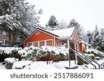 Charming orange house in a snowy winter wonderland. A cozy cottage covered in fresh snow, surrounded by snow-laden trees and shrubs, creates a picturesque winter scene.