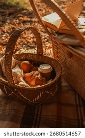A charming picnic setup in the fall, featuring a wicker basket filled with apples, a croissant, and coffee, alongside an open book.