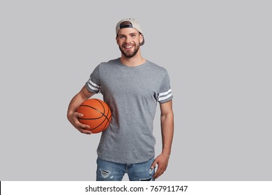 Charming player. Handsome young smiling man carrying a basketball ball and looking at camera while standing against grey background - Powered by Shutterstock