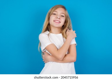 Charming pleased beautiful caucasian little girl wearing white T-shirt over blue background embraces own body, pleasantly feels comfortable poses. Tenderness and self esteem concept - Shutterstock ID 2045715956