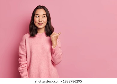 Charming pleased young Asian woman with rouge cheeks points aside on copy space shows advertisement smiles gently wears casual jumper isolated over rosy background. Attention to this promotion - Shutterstock ID 1882986295