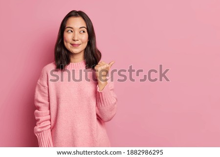 Charming pleased young Asian woman with rouge cheeks points aside on copy space shows advertisement smiles gently wears casual jumper isolated over rosy background. Attention to this promotion