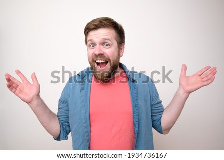 Charming, positive man makes a greeting gesture with arms outstretched to the side, he is smiling and looking at the camera happily. 