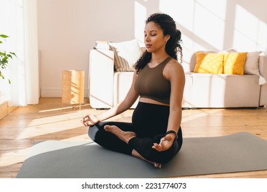 Charming pregnant african american woman in leggings and crop top sitting on floor in living-room in lotus posture with closed eyes, doing mudra sign with fingers, reaching zen, balance and harmony
