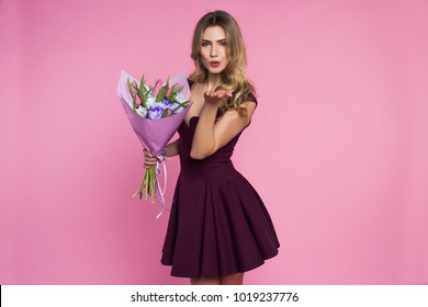 Charming, pretty girl is holding a big tulip bouquet in hands. Lady is sending a vexed kiss. Pink background. Happy woman's day