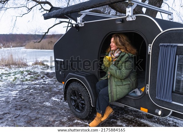 Charming red haired middle aged woman in camper\
trailer alone. Single traveling woman warming up drinking hot\
drink, camper parked under tree to enjoy time and relax at the\
nature. Journey\
concept.