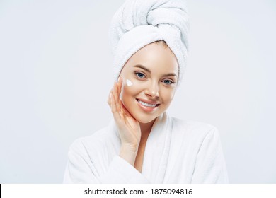 Charming Relaxed Beauty Woman Applies Face Cream, Cares About Complexion, Touches Cheek With Hand, Smiles Gently At Camera, Dressed In Bath Robe, Wrapped Towel On Washed Hair, Isolated On White