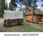 Charming rustic cabin with an old covered wagon in the foreground. The weathered wood and antique wagon evoke a sense of history and nostalgia in this tranquil, picturesque setting.