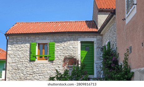 Charming rustic house made of stone with vibrant green wooden shutters and door, red clay tiled roof, flowering plants, and clear blue sky in the background.