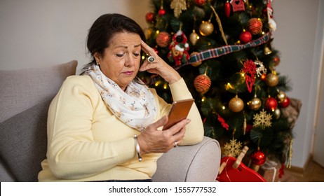 Charming Sad Old Woman Is Sitting On Sofa And Looking On Smartphone. Senior Woman Sitting Alone On Sofa, Christmas Tree In Background. Serious Old Woman Is Expressing Hopeless