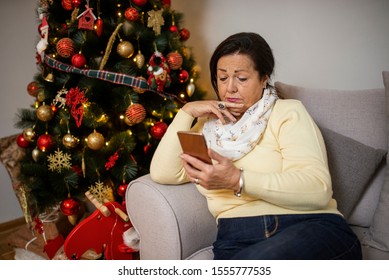 Charming Sad Old Woman Is Sitting On Sofa And Looking On Smartphone. Senior Woman Sitting Alone On Sofa, Christmas Tree In Background. Serious Old Woman Is Expressing Hopeless