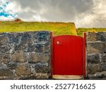 Charming scene of a short red door through a cobblestone wall that leads to red Scandinavian style buildings with natural grass rooftops. Located in Tórshavn, the Capital of the Faroe Islands.