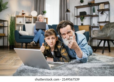 Charming schoolboy playing with grandfather using laptop lying on warm floor in living room while senior grandma rest on sofa in background. Grandparents and grandchild relax at home showing thumb up.