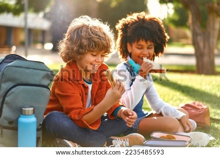 Charming schoolchildren after school resting sitting on the grass in the park. An African-American girl with a Caucasian boy took out lunchboxes with sandwiches from their backpack and eating.