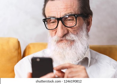 Charming Senior Bearded Man In Glasses Wearing White Shirt Sitting On The Yellow Sofa And Scrolling Facebook