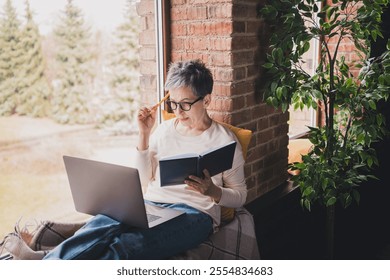 Charming senior woman enjoying leisure time at home, sitting by window with laptop and book. - Powered by Shutterstock