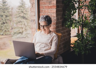 Charming senior woman with short grey hair enjoying leisure time at home, using laptop near window in cozy daylight apartment - Powered by Shutterstock