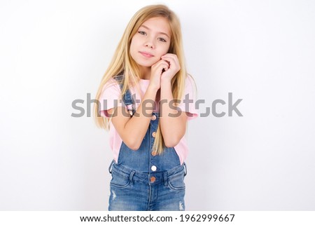Charming serious beautiful caucasian little girl wearing jeans overall over white background keeps hands near face smiles tenderly at camera