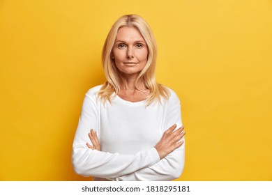 Charming serious middle aged blonde woman stands crossed hands indoor looks directly at camera being confident in future wears casual white jumper poses determined against yellow background.