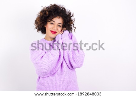 Charming serious Young beautiful Arab woman wearing knitted sweater standing against white background  keeps hands near face smiles tenderly at camera