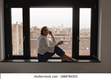 Charming short-haired woman sits on windowsill and enjoys coffee. Lady in jeans and white blouse holds cup on city background.