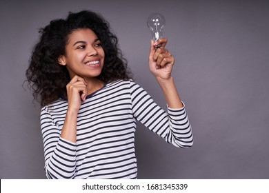 Charming smiling cute young ethnic girl with curly gorgeous hair, in a striped casual jacket, holds a light bulb in her left hand, smiles and looks at her. Concept i have an idea.