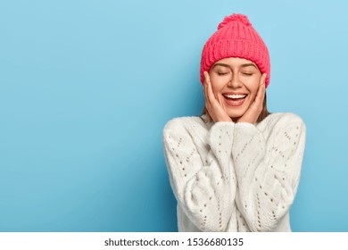 Charming Smiling Feminine Girl Touches Cheeks With Both Hands, Eyes Shut, Toothy Alluring Smile, Wears Winter White Sweater, Poses Against Blue Studio Wall, Has Pure Healthy Skin, Feels Relieved