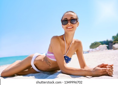 Charming smiling slim woman wearing in bikini is lying on white sand on the sea beach. Woman with sunglasses is sunbathing at heat sunny day at summer vacation