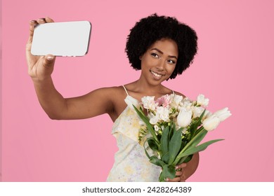 Charming smiling young african american woman with curly hair taking a selfie, holding a smartphone and a bouquet of white and pink tulips on a soft pink background, studio
