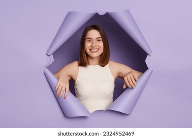 Charming smiling young brunette woman in casual clothing breaking through purple paper hole being in good mood looking with happy face friendly expression. - Shutterstock ID 2324952469