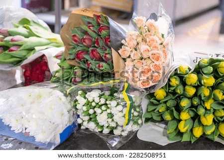 Charming spring yellow and dark red tulips, soft pink roses, delicate white carnations, large white marigolds in transparent packages. The shelf in blurred background.