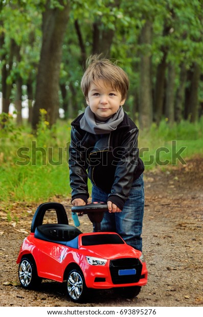 Charming\
stylish brunette boy in a leather jacket and gray scarf on a red\
classic toy car on the road in the\
summer