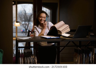 Charming Successful Woman, Confident Sales Manager In Furniture Shop, Interior Designer Standing At Work Desk And Choosing Fabric Swatches For Her Project While Talking On A Mobile Phone With Client