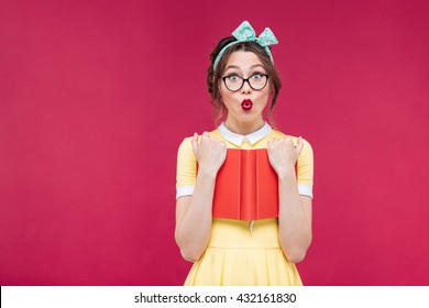 Charming surprised pinup girl in glasses standing and holding red book over pink background