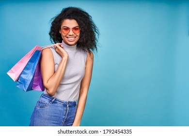Charming surprised young girl with dark skin and curly hair, in red sunglasses, holds a pair of colored shopping packages, posing on a blue background in the studio.