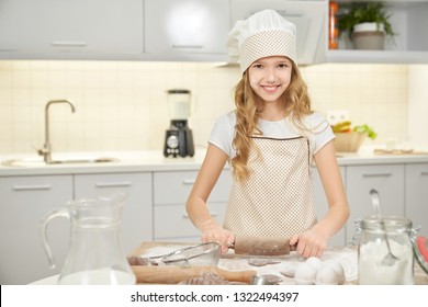 Charming teenager standing on kitchen, holding rolling pin, cooking biscuits. Pretty girl wearing in apron and chef hat looking at camera, smiling and posing. Kid using kitchen tools for pastry.