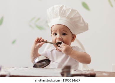 Charming toddler male baby playing as a cooker, dressed in white t-shirt and Chef hat sitting on table with flour spread on cutting board and kitchen utensils