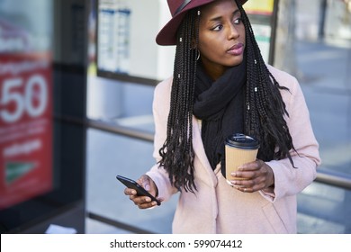 Charming trendy dressed afro american female hipster girl in hat waiting for bus on station in morning with cup of coffee to go using smartphone and 5G connection to monitoring transport through app
