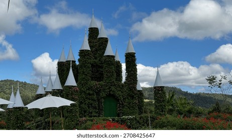 A charming, whimsical castle structure with multiple spires, completely covered in lush green vines and small flowering plants. It stands in a garden with red and green foliage under a vibrant blue 