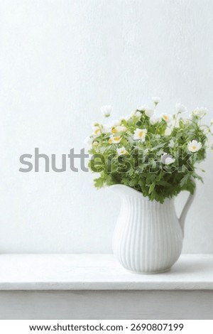 A charming white ceramic pitcher overflows with small white blossoms and green foliage, set against a soft, light background.