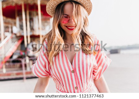 Charming white girl in straw hat laughing on blur city background. Pretty european young lady in striped dress having fun in amusement park.
