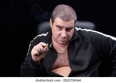 A Charming White Man Smokes A Cigar In A Chair And Looks At The Camera With A Smile From Under His Brows. He Looks Like A Kind Bandit Or A Smiling Russian Mafia,