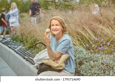 Charming White Senior Lady 62 Years Old Is Walking In The Public Park And Sitting On The Bench Marked For Social Distance During Coronavirus Covid-19 Pandemic With Ice Cream And Smiling.