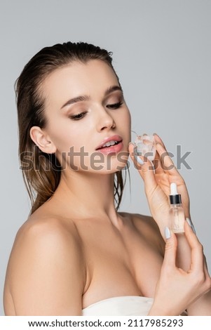 charming woman with bare shoulders, ice cube and cosmetic serum isolated on grey