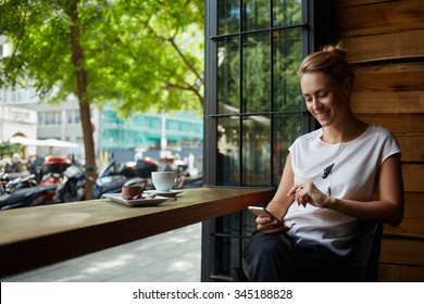 Charming woman with beautiful smile reading good news on mobile phone during rest in coffee shop, happy Caucasian female watching her photo on cell telephone while relaxing in cafe during free time 