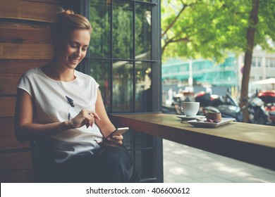 Charming Woman With Beautiful Smile Reading Good News On Mobile Phone During Rest In Coffee Shop, Happy Caucasian Female Watching Her Photo On Cell Telephone While Relaxing In Cafe During Free Time 